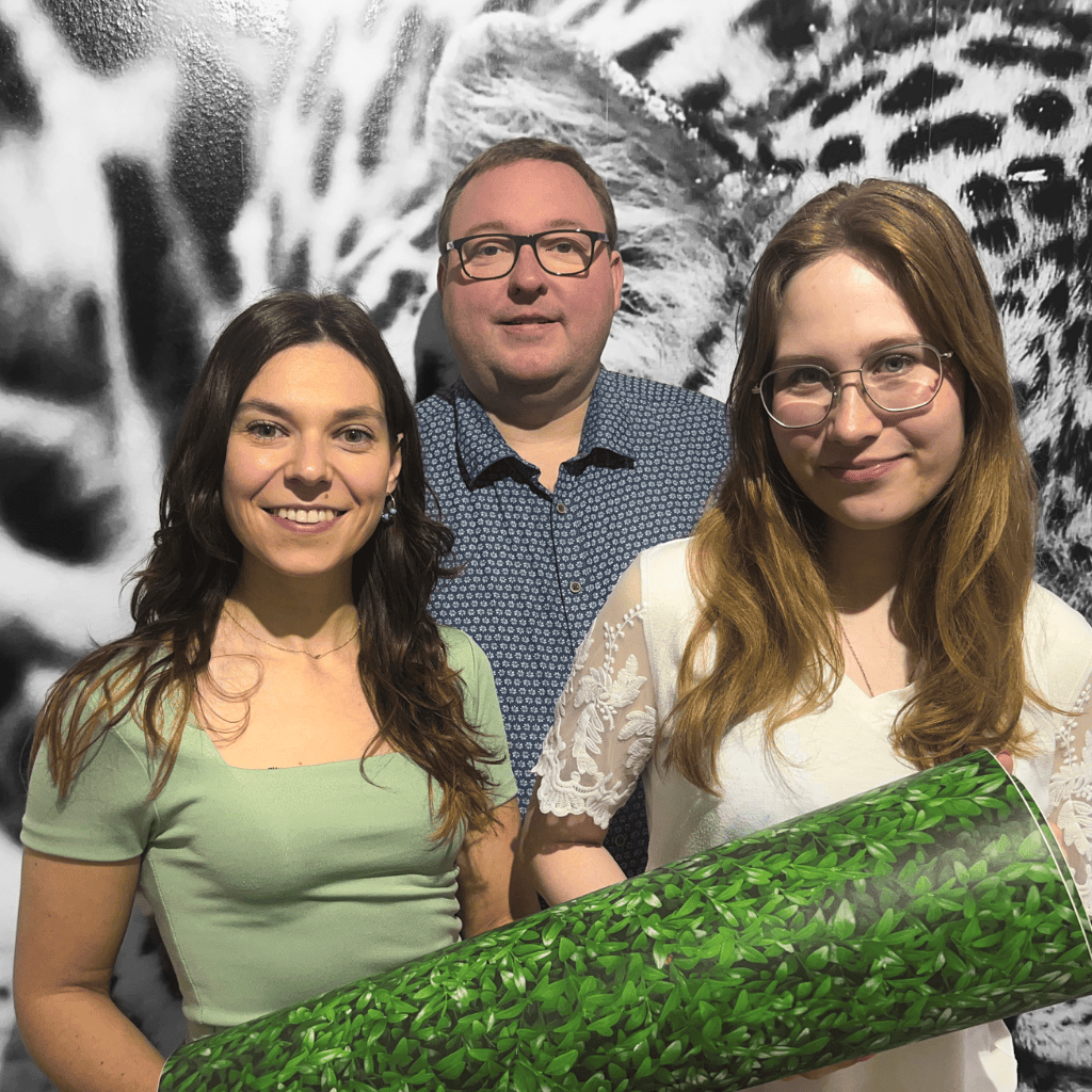Drei Teammitglieder stehen vor einer schwarz-weißen Leopardenmuster-Fototapete, eine Person hält eine grüne Tapetenrolle in der Hand
